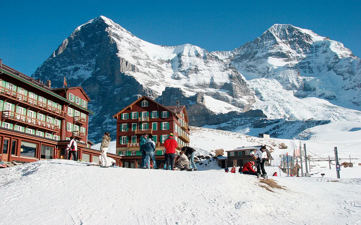 Hotel am Fuße schneebedeckter Berge mit Menschen vor dem Gebäude.