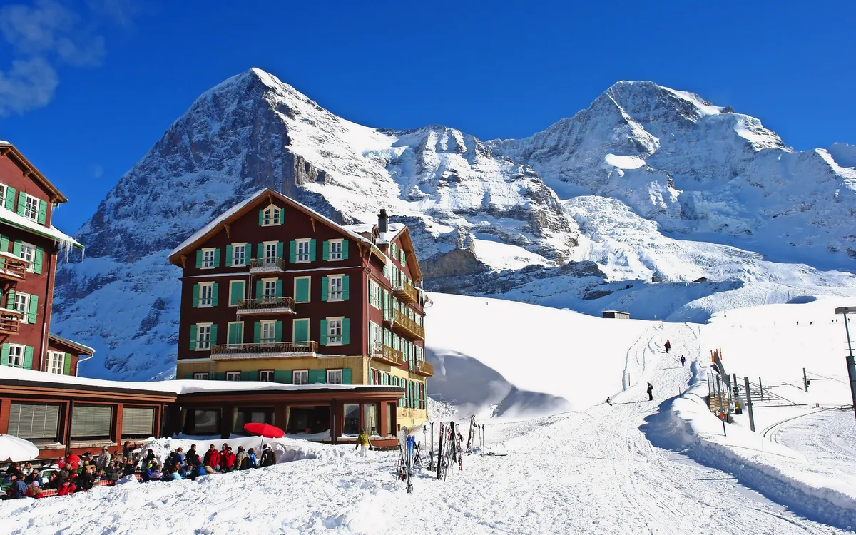 Berge im Schnee mit Hotel und Skifahrern, strahlend blauer Himmel im Hintergrund.