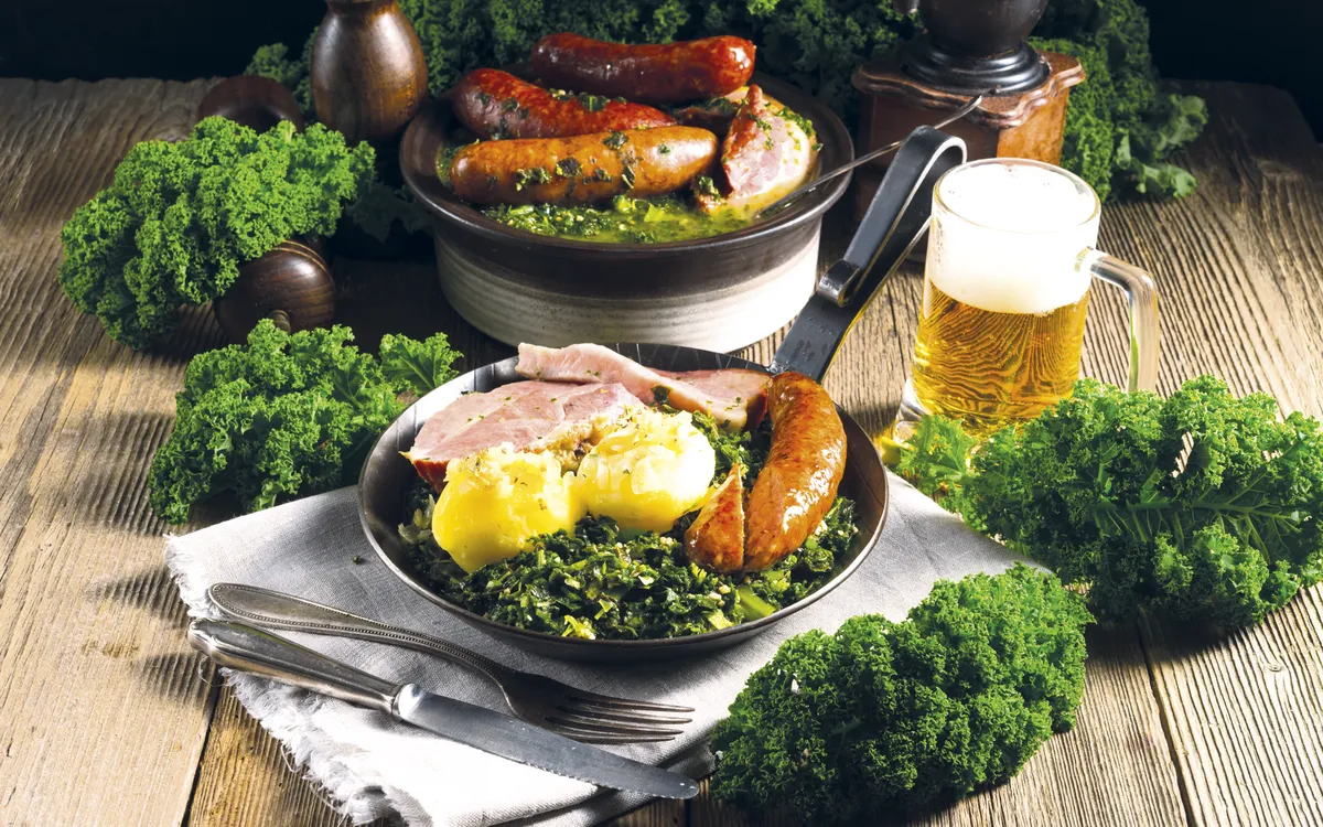 Grünkohlgericht mit Kassler, Pinkel, Kartoffeln und Bier auf Holztisch.