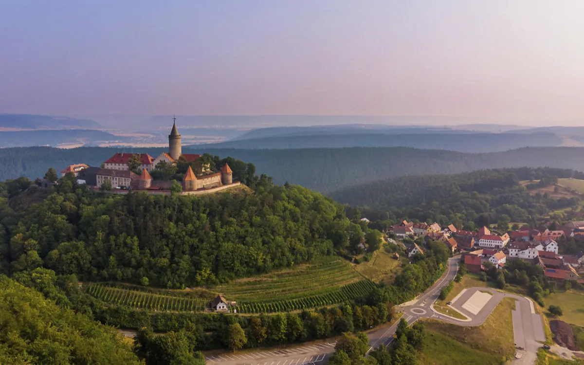Burg auf bewaldetem Hügel mit umliegender Landschaft und Dorf im Hintergrund.