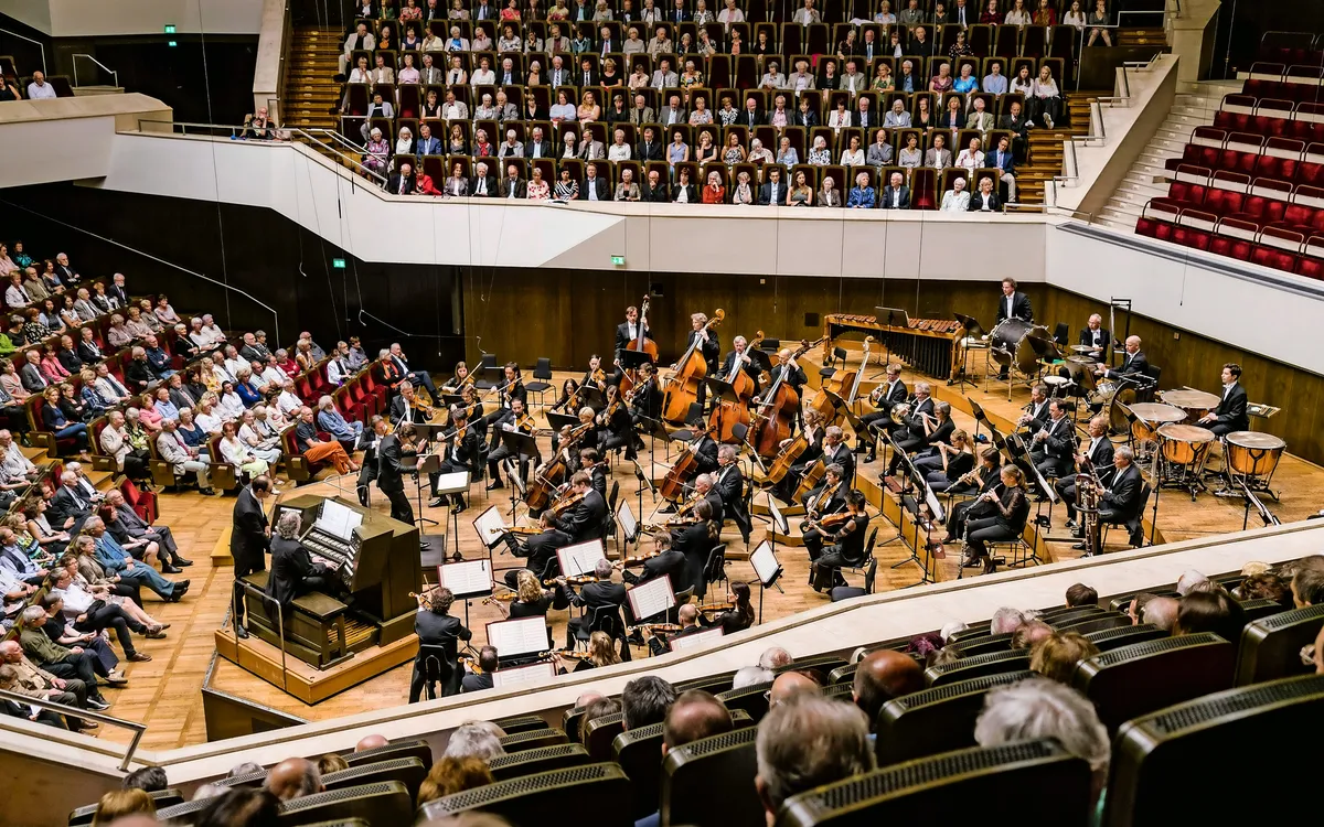 Orchester spielt in einem Konzertsaal, Publikum sitzt gespannt.