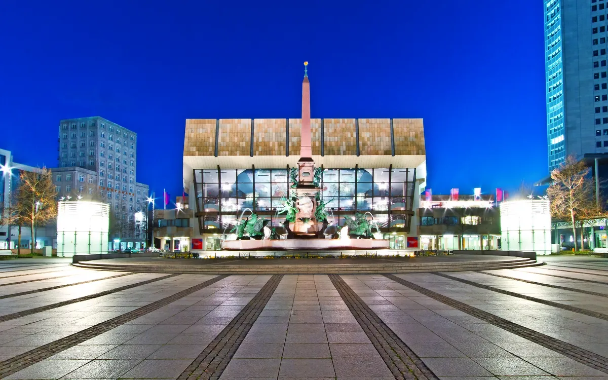 Gebäude mit moderner Glasfassade bei Nacht, im Vordergrund ein beleuchteter Brunnen.
