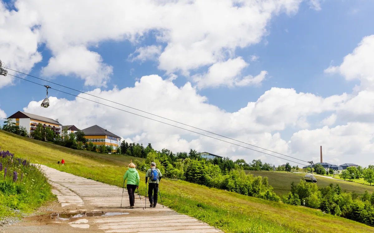 Zwei Personen wandern einen Pfad entlang, Seilbahn und Landschaft im Hintergrund.