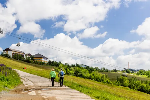 Zwei Personen wandern einen Pfad entlang, Seilbahn und Landschaft im Hintergrund.