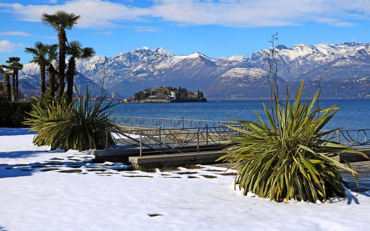 Schneebedeckter Garten mit Palmen, Hintergrund Berge und See mit Schloss.