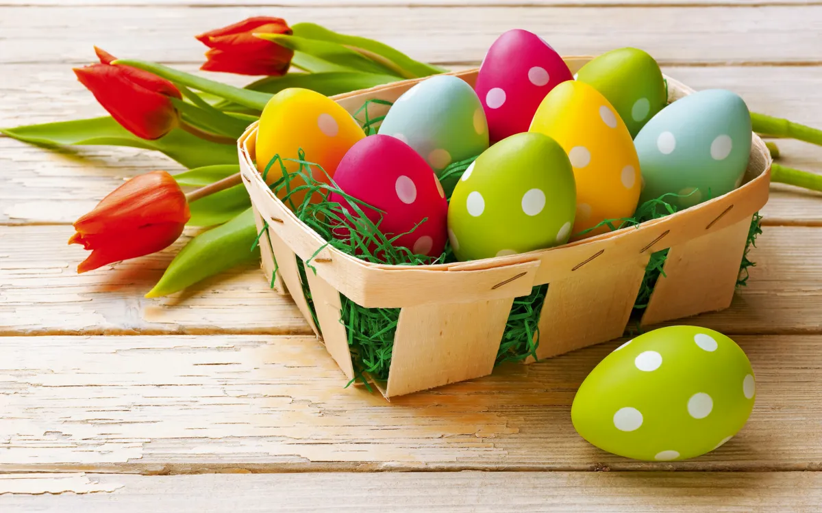 Osterkorb mit bunten, gepunkteten Eiern auf einem Holztisch, daneben liegen rote Tulpen.