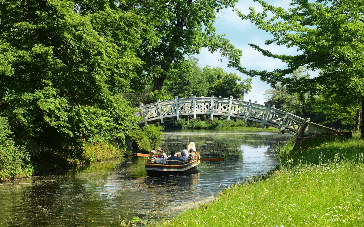 Boot auf einem Fluss unter einer Holzbrücke, umgeben von Bäumen.