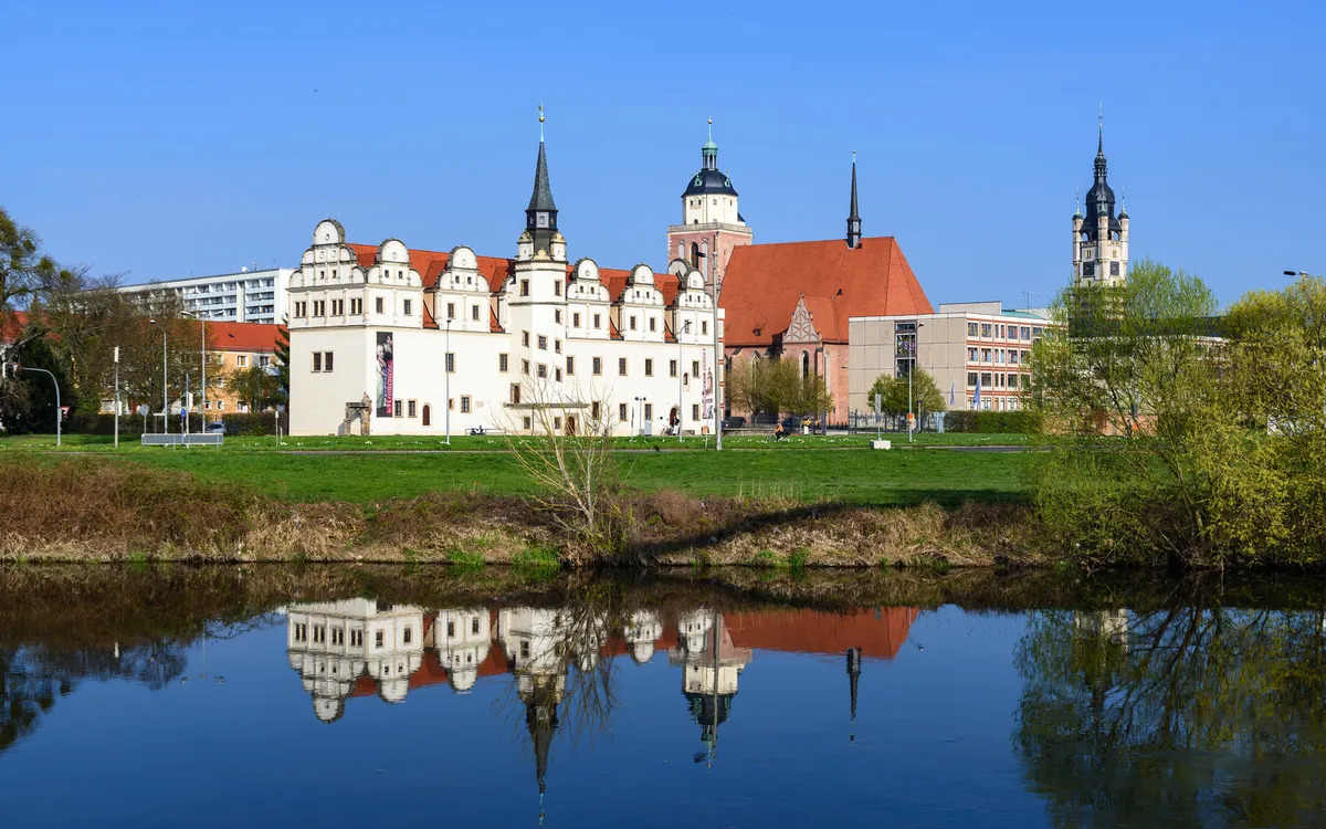Schloss und Kirche spiegeln sich in einem Fluss bei klarem Himmel.