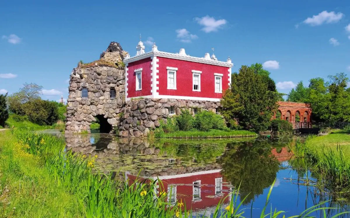 Ein rotes Gebäude mit Steinruine am Teich, umgeben von grüner Vegetation.