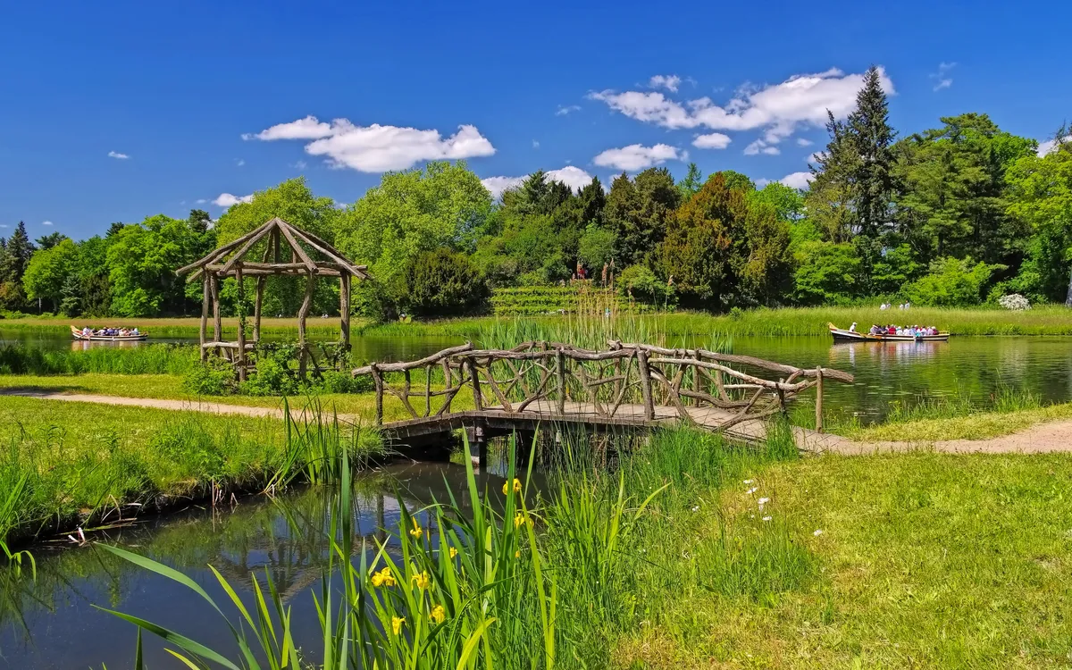 Grüne Parklandschaft mit Holzbrücke, Pavillon und Booten im Wasser.