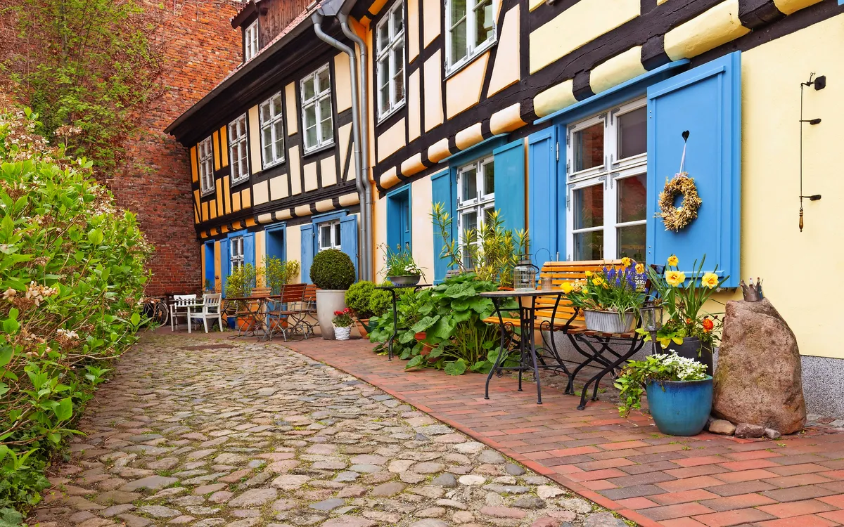 Fachwerkhaus mit blauen Fensterläden, Blumendeko und Kopfsteinpflaster in schmalem Innenhof.