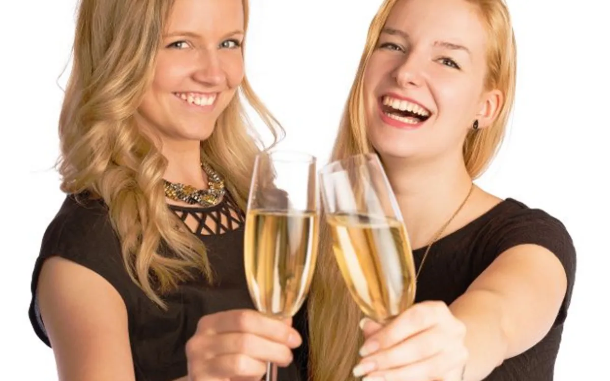 Zwei Frauen in schwarzer Kleidung stoßen mit gefüllten Sektgläsern an und lächeln in die Kamera.