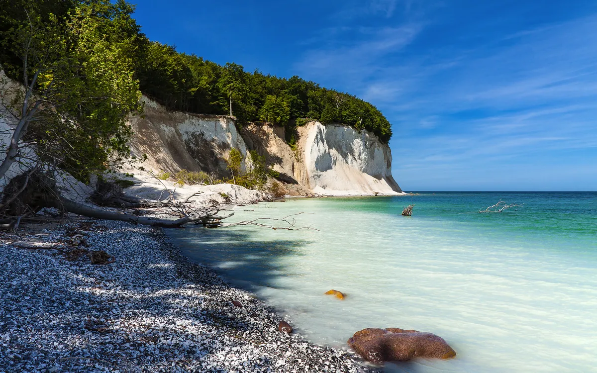 Kreidefelsen an einer Küste unter blauem Himmel, mit Kiesstrand und Bäumen.
