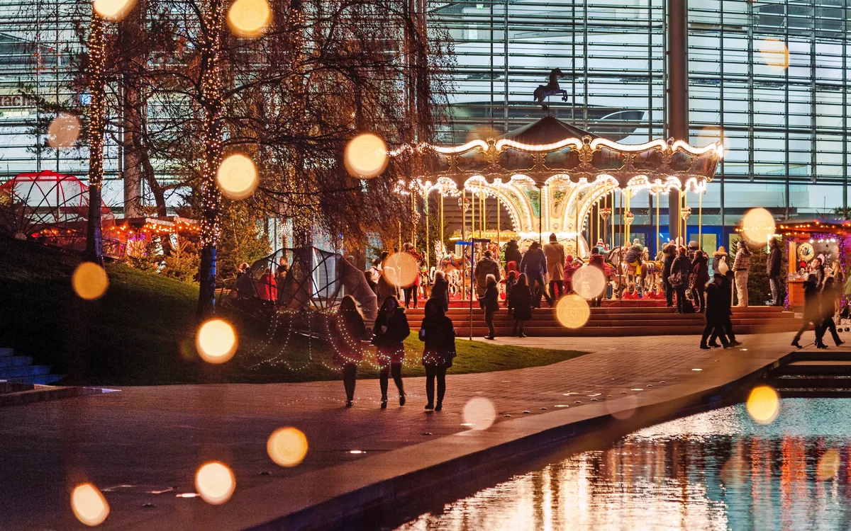 Menschen spazieren bei Nacht an einem beleuchteten Karussell und Teich vorbei.