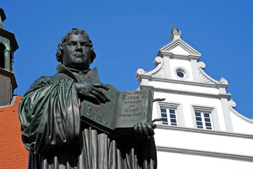 Statue eines Mannes mit aufgeschlagenem Buch vor blauem Himmel und historischem Gebäude.