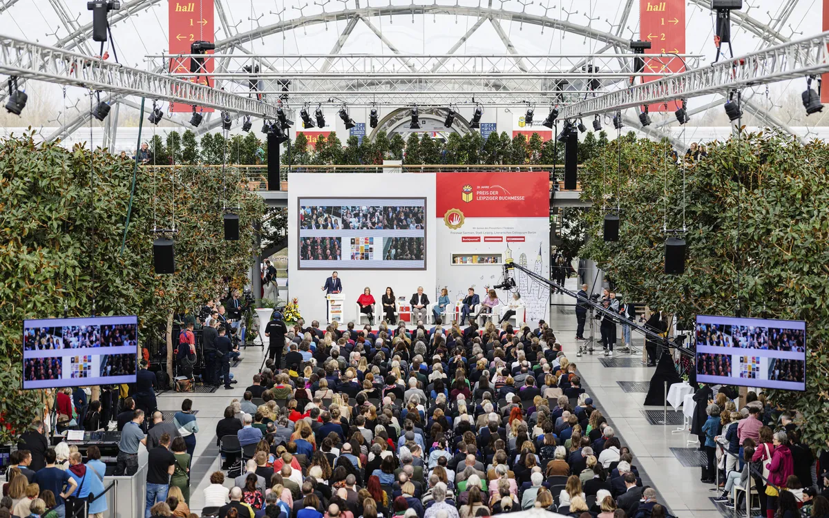 Menschen auf der Leipziger Buchmesse vor einer Bühne mit Rednern und Bildschirmen.