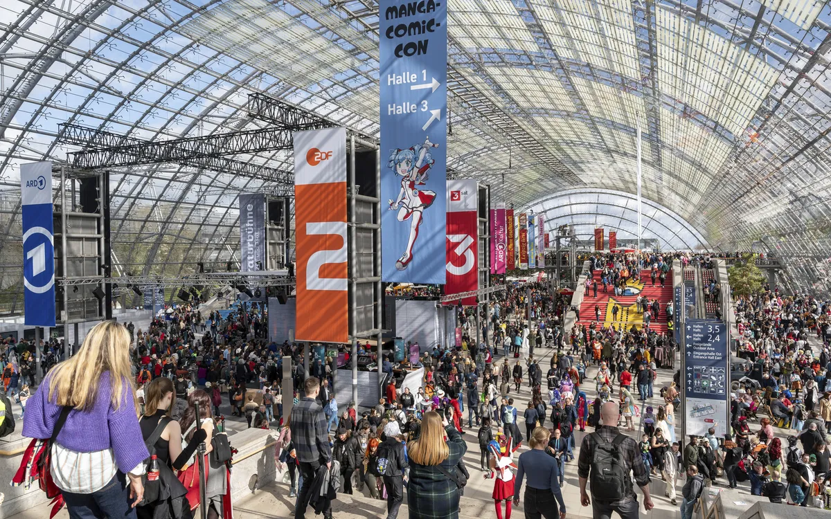 Menschenmenge in einer großen Messehalle mit bunten Bannern und Ständen.