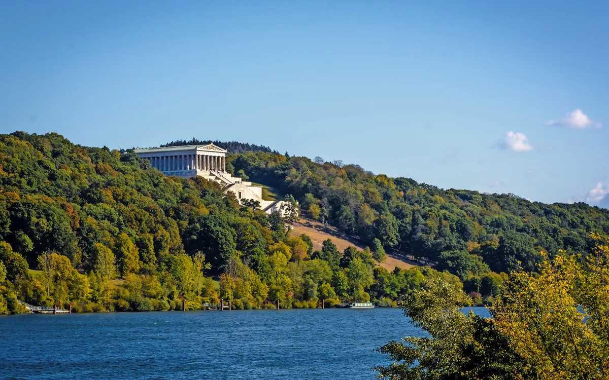 Gebäude auf bewaldetem Hügel am Flussufer unter klarem Himmel.
