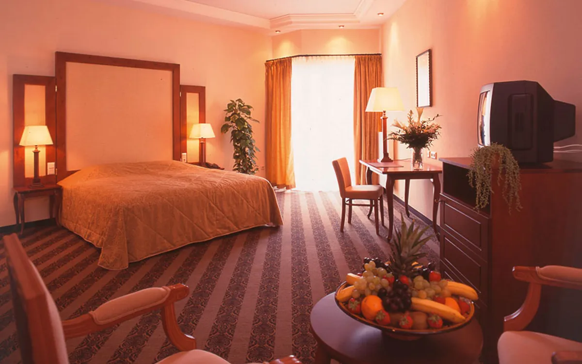 Hotelzimmer mit Doppelbett, Schreibtisch und Fernseher, warm beleuchtet, mit Früchteschale.
