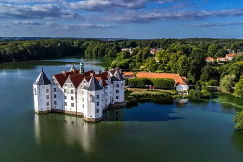Weißes Schloss auf einer Insel in einem See, umgeben von Wald und blauen Himmel.