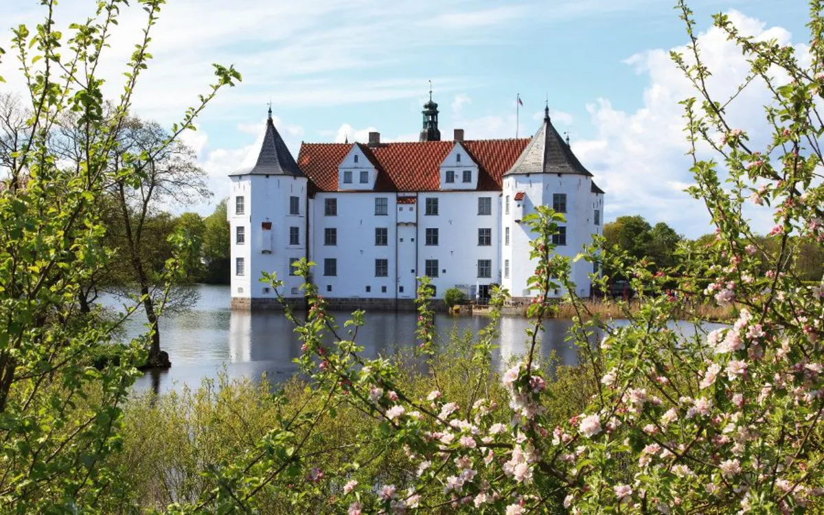 Weißes Schloss mit Türmen hinter blühenden Bäumen, umgeben von Wasser.