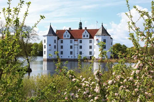 Weißes Schloss mit Türmen hinter blühenden Bäumen, umgeben von Wasser.