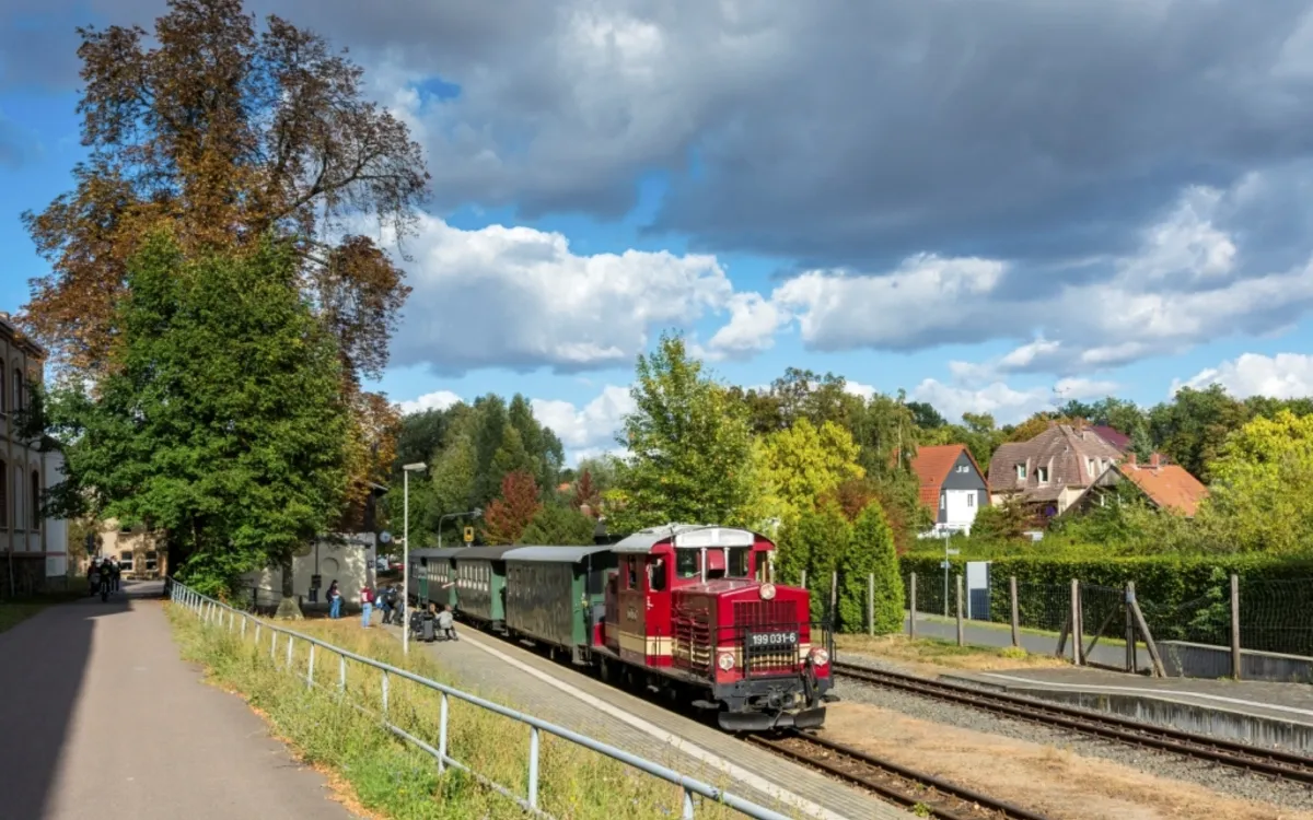 Roter Zug der Döllnitzbahn fährt bei Sonnenschein durch ländliche Umgebung.