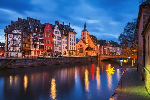 Fachwerkhäuser und eine Kirche am Flussufer bei Nacht, mit festlicher Beleuchtung.