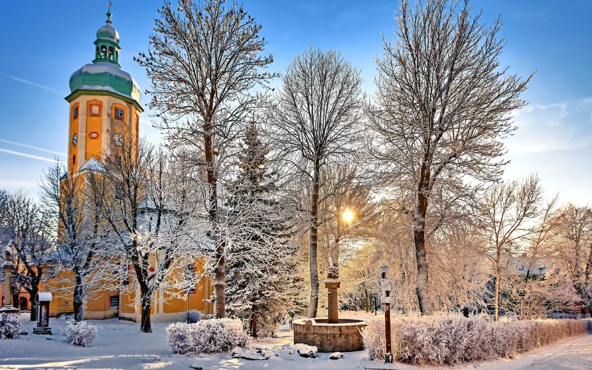 Verschneiter Park mit Bäumen und gelber Kirche im Hintergrund.