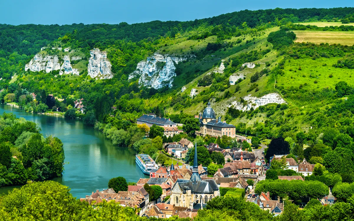Landschaft mit Dorf, Fluss und grünen Hügeln im Hintergrund.