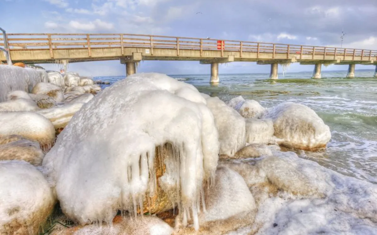 Verschneite Felsen am Strand mit einer Brücke im Hintergrund bei klarem Himmel und leicht bewölkt.