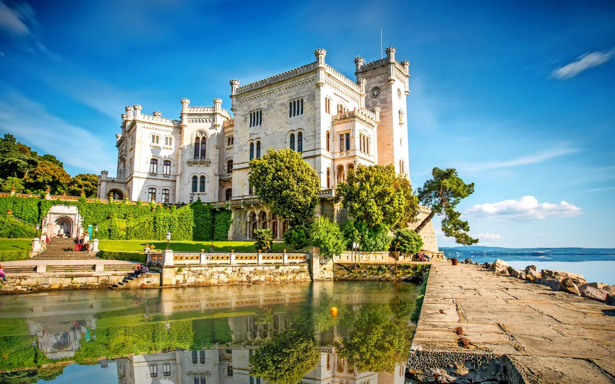 Historisches Schloss am Ufer mit blauem Himmel und reflektierendem Wasser.
