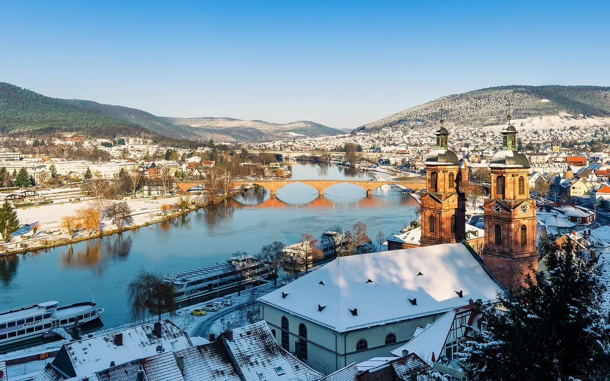 Verschneite Stadtlandschaft mit Fluss und Kirche im Vordergrund.