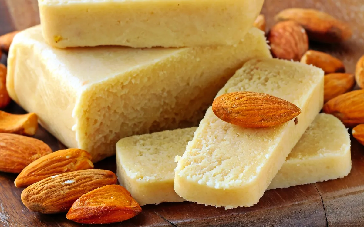 Stücke von Marzipan auf einem Holzbrett, umgeben von Mandeln.