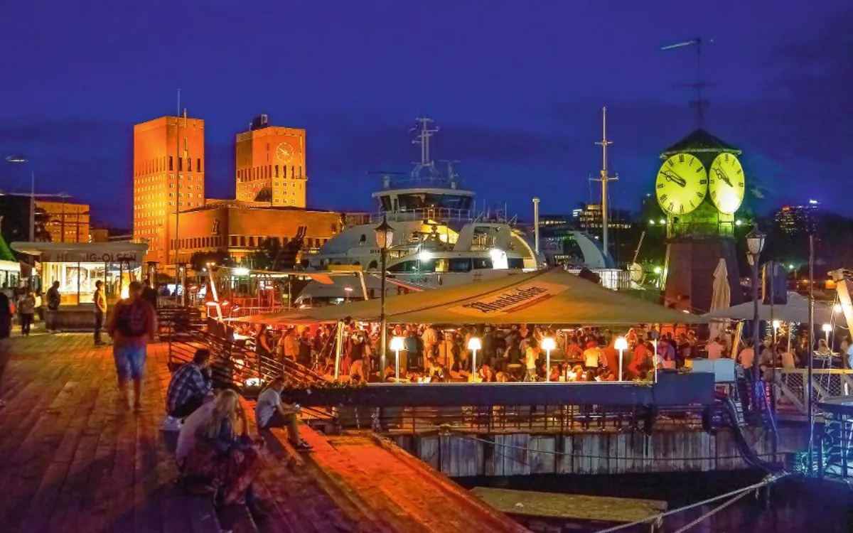 Osloer Rathaus bei Nacht, beleuchtete Uferpromenade mit Menschen und Booten im Vordergrund.