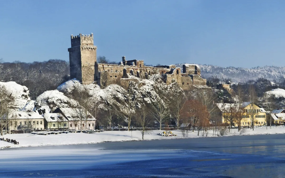 Schneebedeckte Burg über einem Fluss, umgeben von Häusern und Bäumen im Winter.