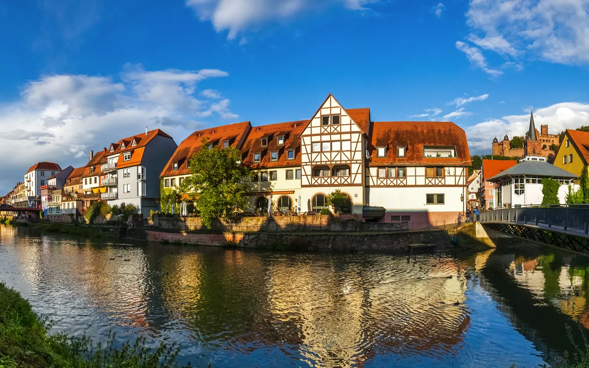 Fachwerkhäuser an einem Fluss bei sonnigem Wetter, gesäumt von Bäumen und blauem Himmel.