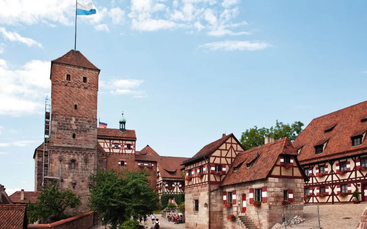 Burg mit Fachwerkgebäuden und blauer Fahne am Turm bei klarem Himmel.