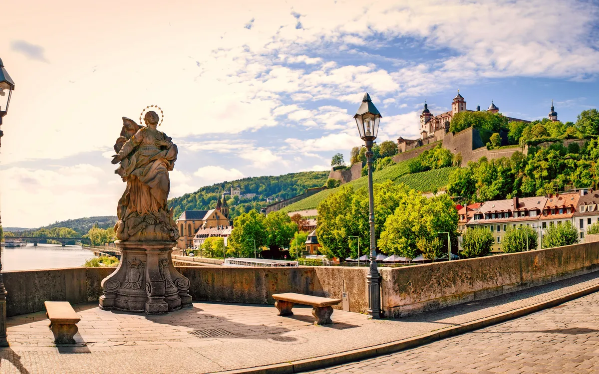 Statue auf einer Brücke mit Festung und Fluss im Hintergrund bei Sonnenschein.