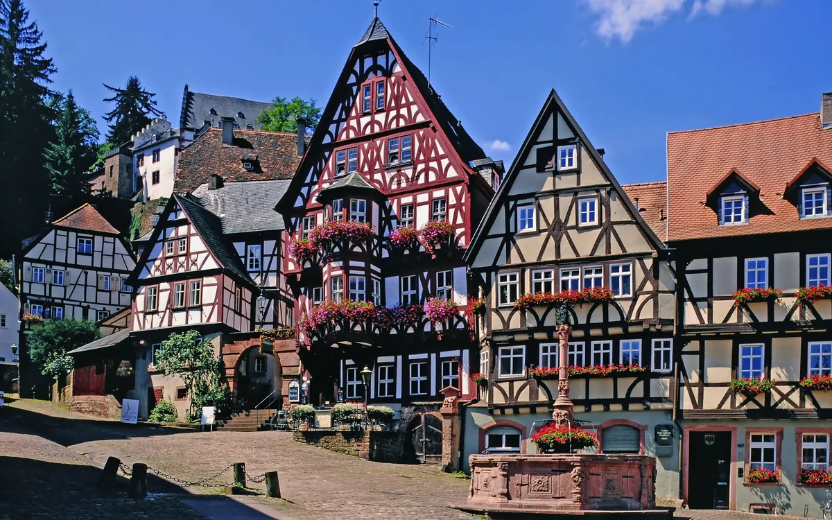 Fachwerkhäuser auf einem gepflasterten Platz mit Blumen und einem Brunnen bei Sonnenschein.