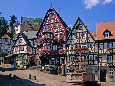 Fachwerkhäuser auf einem gepflasterten Platz mit Blumen und einem Brunnen bei Sonnenschein.