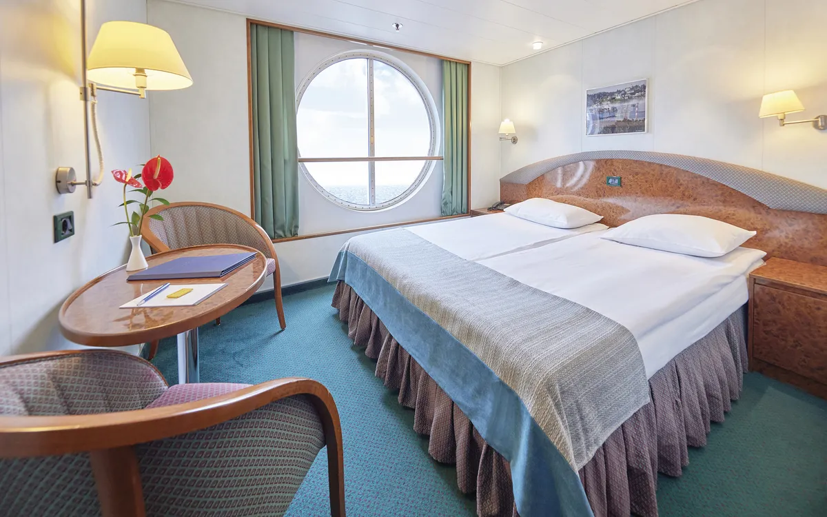 MS Hamburg Außenkabine mit Doppelbett und Sitzbereich, großer runder Fensterblick.