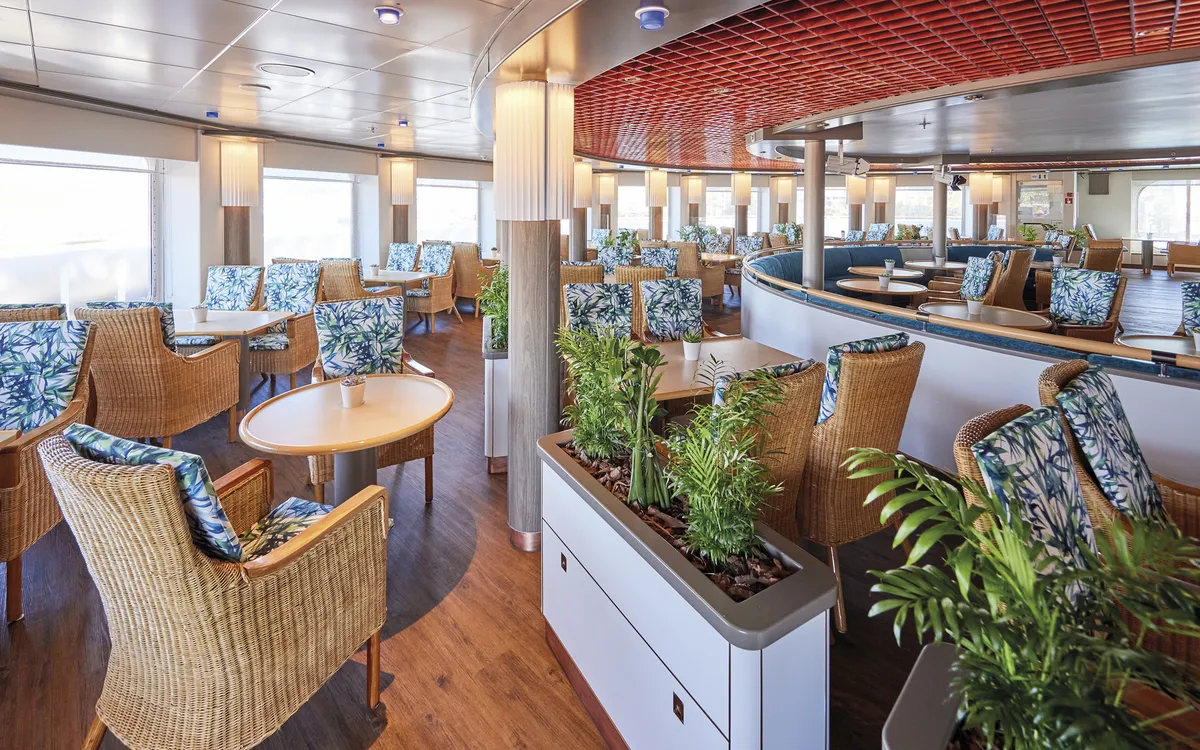 Restaurant im Inneren des Schiffes MS Hamburg mit Tischen und geflochtenen Stühlen.