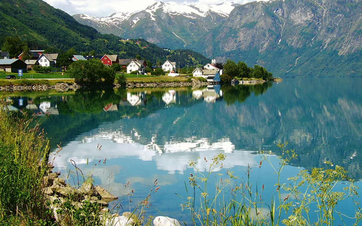 Kleiner Ort am See mit Holzhäusern, Spiegelung, Berge, schneebedeckte Gipfel, Gräser im Vordergrund.