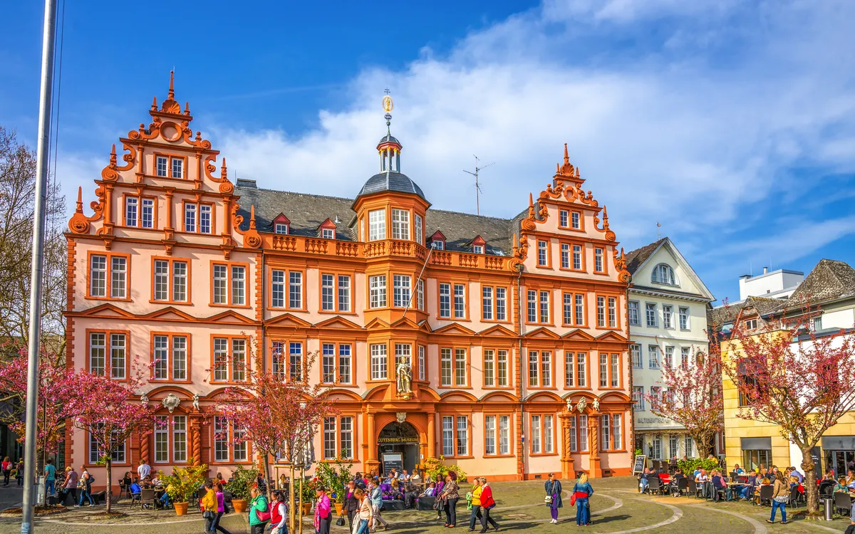Historisches Gebäude mit roten Fensterläden und blühenden Bäumen davor.