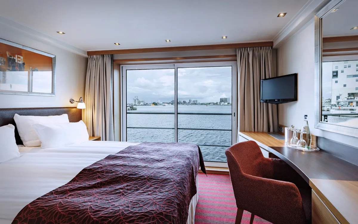 Heller Kabinenraum mit Doppelbett, Schreibtisch, Fernseher und Blick aufs Wasser.