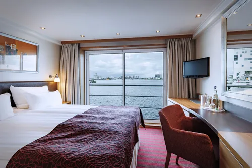 Heller Kabinenraum mit Doppelbett, Schreibtisch, Fernseher und Blick aufs Wasser.