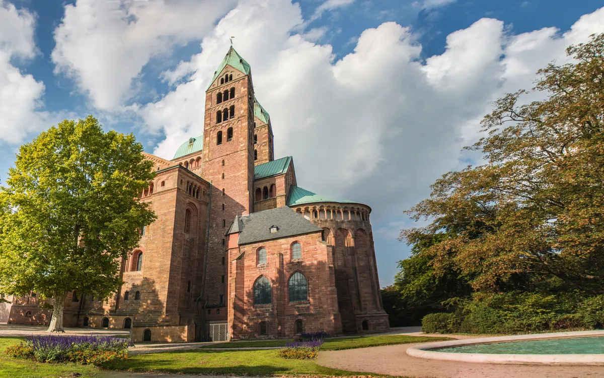 Historisches Gebäude mit Türmen, umgeben von Bäumen und einem Brunnen im Vordergrund.