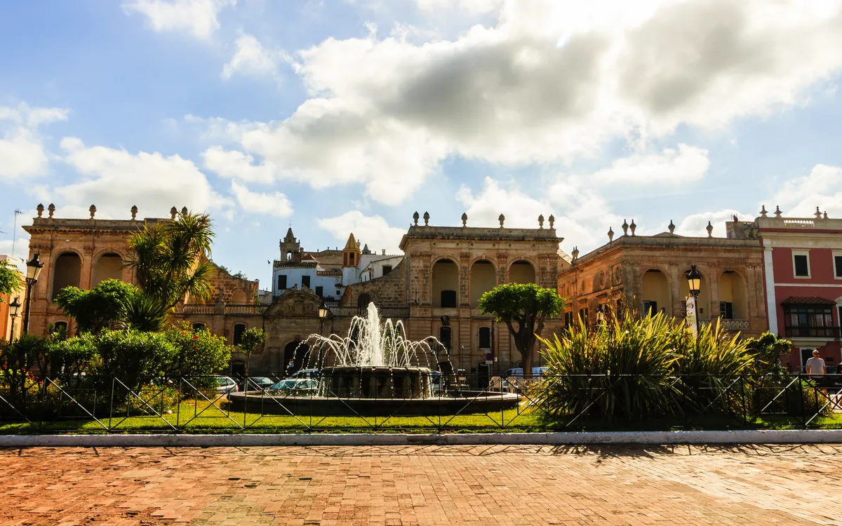 Historische Gebäude und Brunnen in Ciutadella, Menorca, unter bewölktem Himmel.