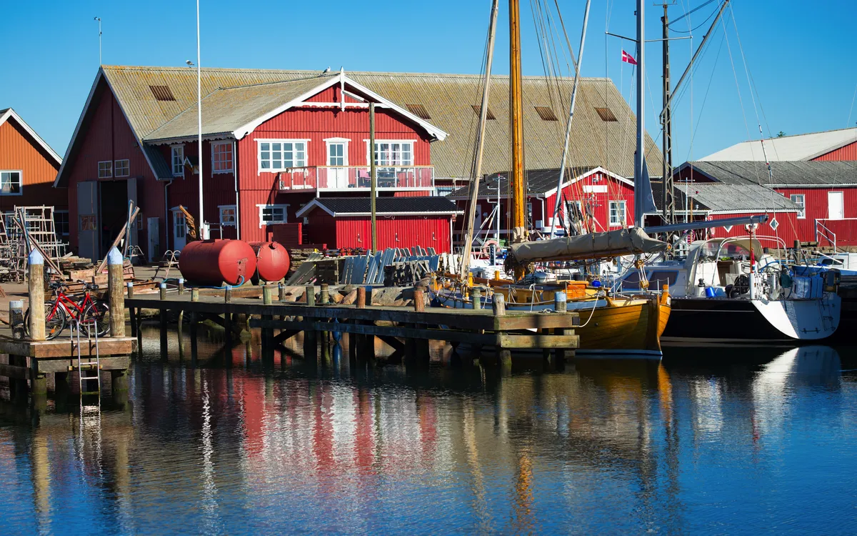 Kleiner Hafen mit roten Holzhäusern und Segelbooten an einem sonnigen Tag.